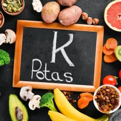 Tablica z napisem "Potas" otoczona produktami bogatymi w ten pierwiastek, jak banany, ziemniaki, pomidory, awokado, brokuły, rodzynki, morele, kiwi, migdały, pestki dyni, orzechy i fasola. Pomaga to zapobiegać niedoborowi potasu.