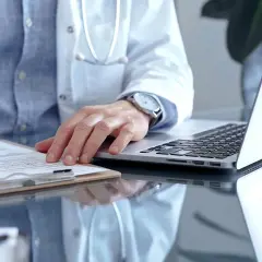 Lekarz w białym kitlu pisze na kartce, obok laptop. Może to być dokumentacja medyczna, np. l4 od psychiatry.
