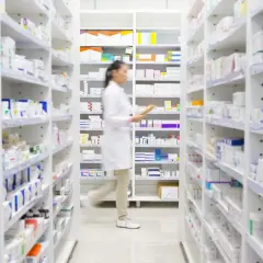 Farmaceutka w białym fartuchu przegląda półki z lekami, szukając np. Klacid.