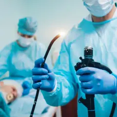 Lekarz w niebieskich rękawiczkach trzyma endoskop. W tle pacjent i inny medyk. Procedura, jak ile trwa gastroskopia, jest krótka.