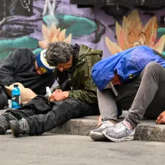 Trzech mężczyzn siedzi na chodniku, pogrążonych w apatii. Obok leży niebieska butelka, a w tle kolorowy mural. To obraz nędzy, gdzie fentanyl narkotyk niszczy życie.