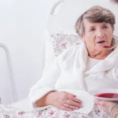 Starsza kobieta w łóżku, karmiona łyżką zupy. Obok stojak rehabilitacyjny. Ocena stanu pacjenta według skali Barthel.