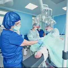 Anestezjolog w niebieskim uniformie przygotowuje pacjenta do zabiegu. W tle zespół medyczny.