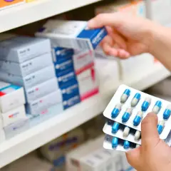Ręka wybiera z półki lek, trzymając opakowania z kapsułkami. Może to być antybiotyk 3 dniowy.