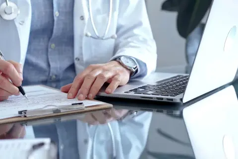 Lekarz w białym kitlu pisze na kartce, obok laptop. Może to być dokumentacja medyczna, np. l4 od psychiatry.