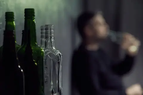 Puste butelki po alkoholu, w tym zielone i przezroczyste, stoją w rzędzie. W tle niewyraźna postać mężczyzny pijącego z butelki, symbolizująca problem z etanolem.