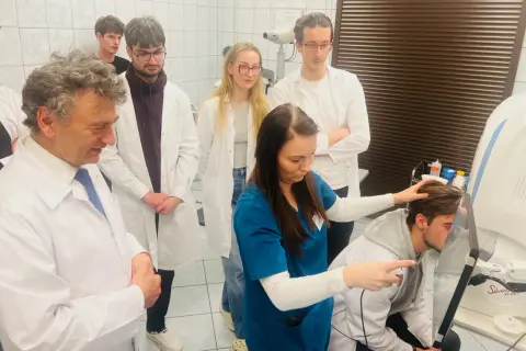 Grupa lekarzy i studentów w białych fartuchach obserwuje badanie pacjenta na nowoczesnym sprzęcie medycznym.