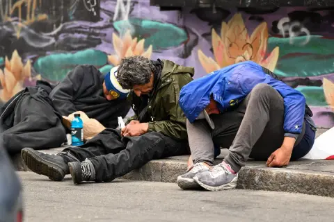 Trzech mężczyzn siedzi na chodniku, pogrążonych w apatii. Obok leży niebieska butelka, a w tle kolorowy mural. To obraz nędzy, gdzie fentanyl narkotyk niszczy życie.