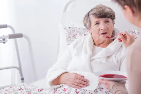 Starsza kobieta w łóżku, karmiona łyżką zupy. Obok stojak rehabilitacyjny. Ocena stanu pacjenta według skali Barthel.