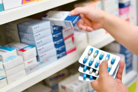 Ręka wybiera z półki lek, trzymając opakowania z kapsułkami. Może to być antybiotyk 3 dniowy.