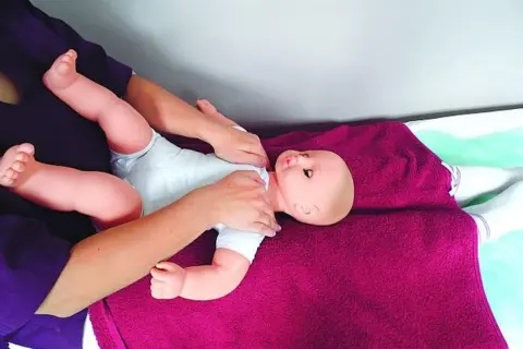Rączki delikatnie masują niemowlęcą laleczkę, ćwicząc masaż Shantala. Dziecko leży na fioletowym kocyku.
