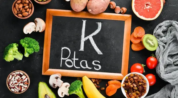 Tablica z napisem "Potas" otoczona produktami bogatymi w ten pierwiastek, jak banany, ziemniaki, pomidory, awokado, brokuły, rodzynki, morele, kiwi, migdały, pestki dyni, orzechy i fasola. Pomaga to zapobiegać niedoborowi potasu.
