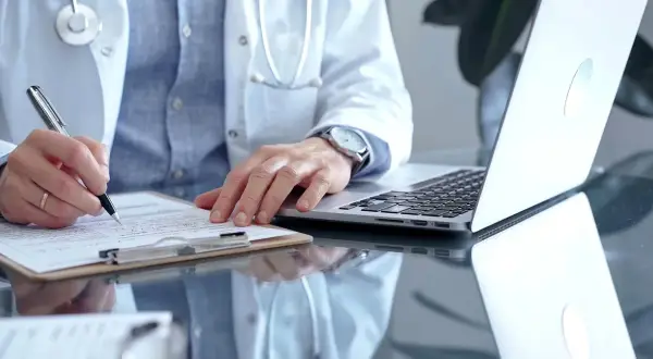 Lekarz w białym kitlu pisze na kartce, obok laptop. Może to być dokumentacja medyczna, np. l4 od psychiatry.