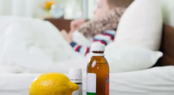 Cytryna, syrop i leki na przeziębienie dla dziecka leżą obok łóżka, gdzie odpoczywa chory maluch.