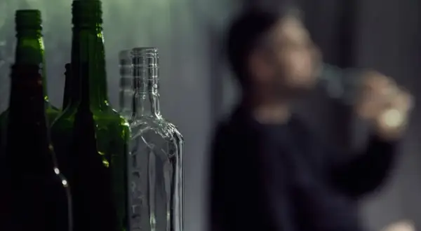 Puste butelki po alkoholu, w tym zielone i przezroczyste, stoją w rzędzie. W tle niewyraźna postać mężczyzny pijącego z butelki, symbolizująca problem z etanolem.