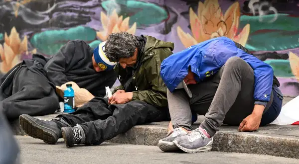 Trzech mężczyzn siedzi na chodniku, pogrążonych w apatii. Obok leży niebieska butelka, a w tle kolorowy mural. To obraz nędzy, gdzie fentanyl narkotyk niszczy życie.