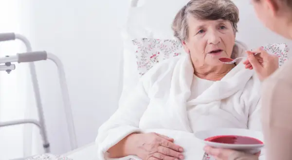 Starsza kobieta w łóżku, karmiona łyżką zupy. Obok stojak rehabilitacyjny. Ocena stanu pacjenta według skali Barthel.