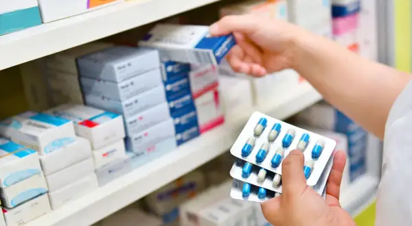 Ręka wybiera z półki lek, trzymając opakowania z kapsułkami. Może to być antybiotyk 3 dniowy.