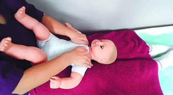 Rączki delikatnie masują niemowlęcą laleczkę, ćwicząc masaż Shantala. Dziecko leży na fioletowym kocyku.