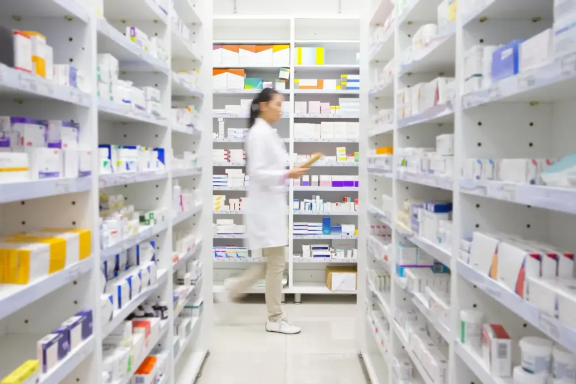 Farmaceutka w białym fartuchu przegląda półki z lekami, szukając np. Klacid.