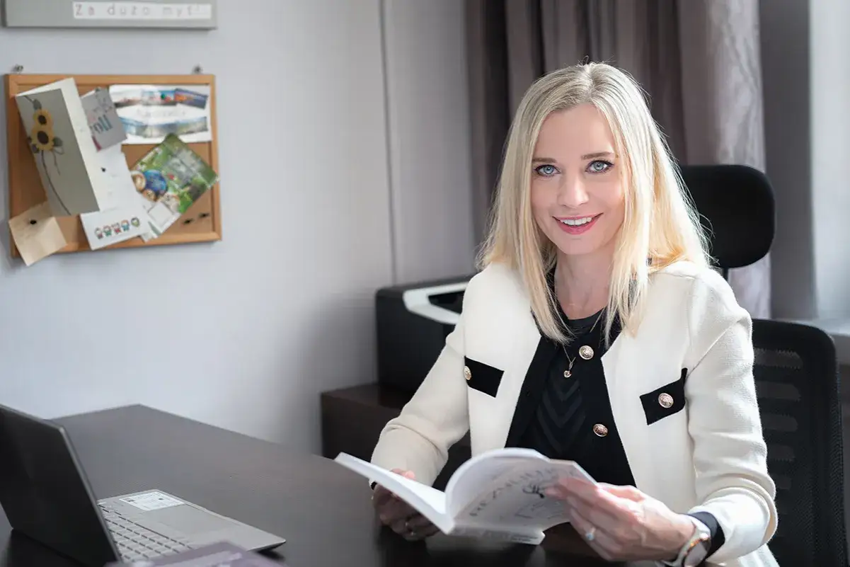 Uśmiechnięta kobieta z blond włosami czyta książkę. W tle tablica z inspiracjami, idealna do planowania terapii behawioralnej.