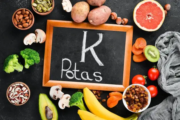Tablica z napisem "Potas" otoczona produktami bogatymi w ten pierwiastek, jak banany, ziemniaki, pomidory, awokado, brokuły, rodzynki, morele, kiwi, migdały, pestki dyni, orzechy i fasola. Pomaga to zapobiegać niedoborowi potasu.