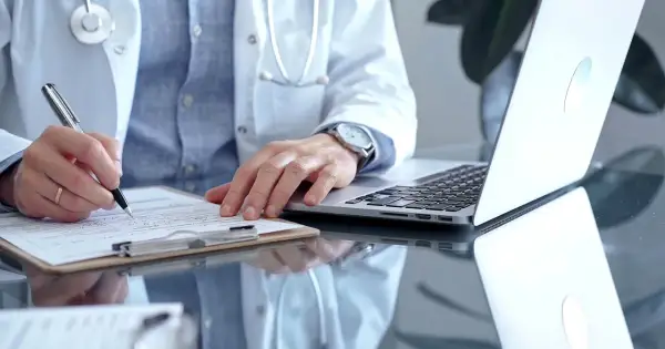 Lekarz w białym kitlu pisze na kartce, obok laptop. Może to być dokumentacja medyczna, np. l4 od psychiatry.