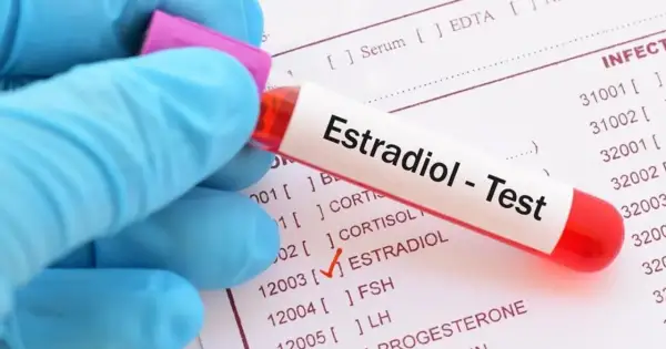 Ręka w niebieskiej rękawiczce trzyma probówkę z napisem "Estradiol - Test". Na papierze obok zaznaczono "ESTRADIOL".