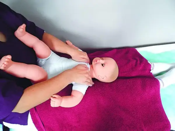 Rączki delikatnie masują niemowlęcą laleczkę, ćwicząc masaż Shantala. Dziecko leży na fioletowym kocyku.